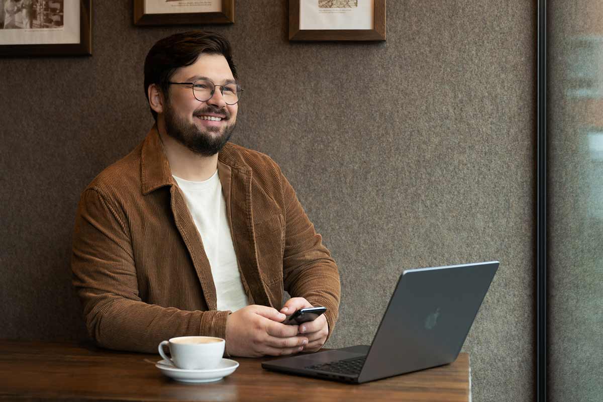 Mann sitzt lächelnd im Café und hat sein Handy in der Hand." title="Mann im Café denkt über den Arbeitgebermarkt nach