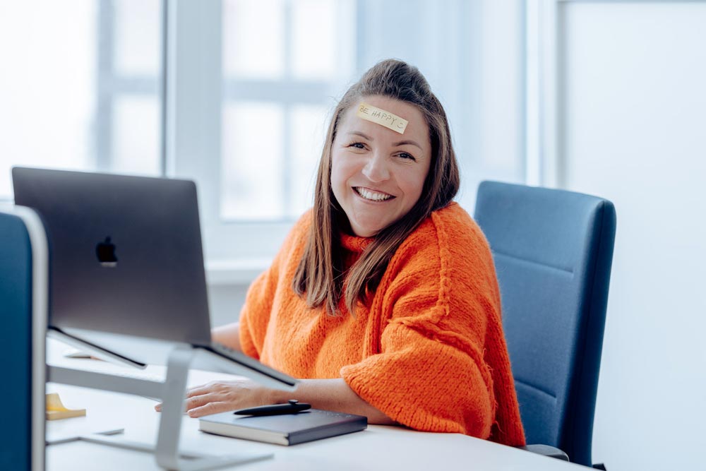 Lächelnde Frau mit orangenen Pulli sitzt am Schreibtisch und hat einen Zettel mit der Aufschrift "BE HAPPY" auf der Stirn kleben" title="Conny „BE HAPPY“