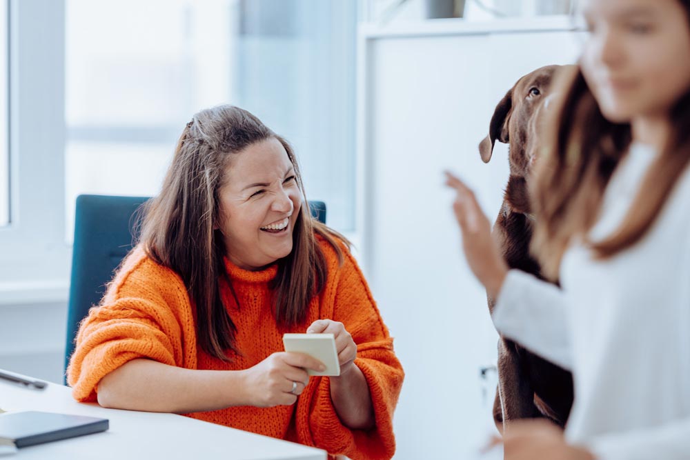 Lachende Frau mit orangenen Pulli sitzt am Schreibtisch und hält Notizzettel in der Hand" title="Conny im Büro