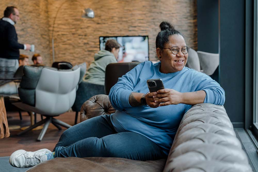 lächelnde Frau in blauem Pullover sitzt mit Handy in der Hand auf einer Couch und schaut aus dem Fenster." title="Tina Kambobe auf einer Couch mit Handy