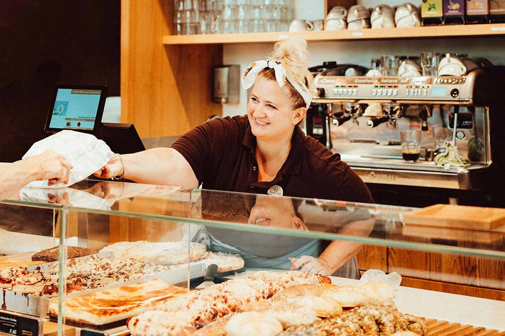 lächelnde Bäckersfrau reicht hinter den Tresen dem Gast die Tüte" title="e2n Software Branche Bäckerei