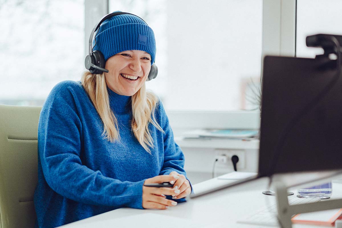 blonde Frau mit blauem Outfit sitzt lachend am Schreibtisch: intrinsische Motivation fördern" title="Luise am Schreibtisch lachend