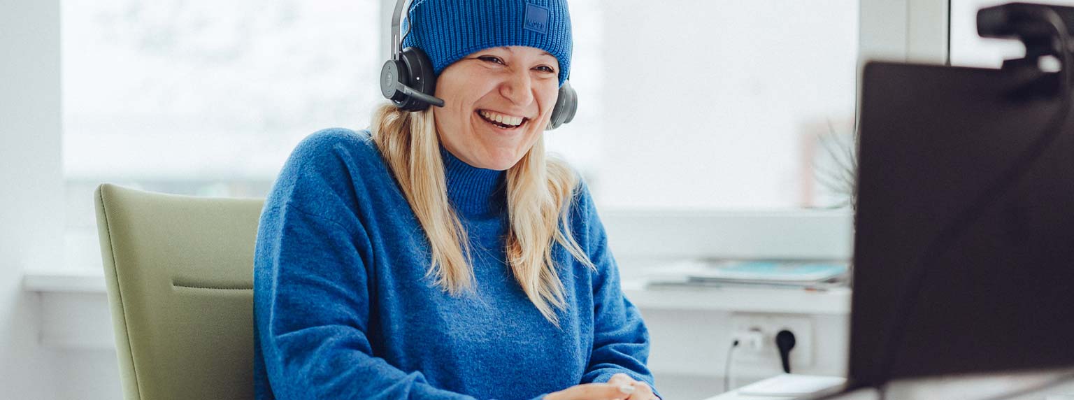 blonde Frau mit blauem Outfit sitzt lachend am Schreibtisch: intrinsische Motivation fördern" title="Luise am Schreibtisch