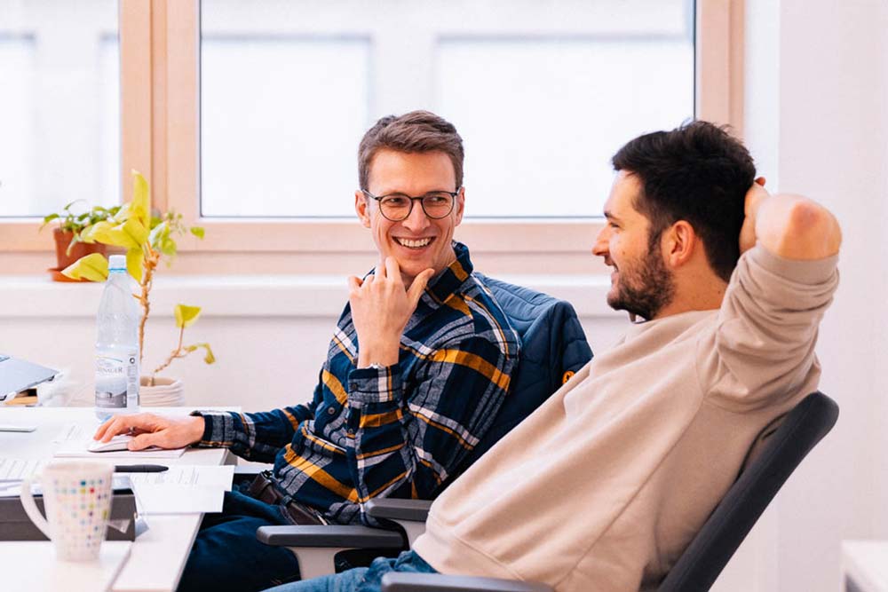 zwei Männer sitzen lächelnd am Schreibtisch" title="Paul und Robert am Schreibtisch