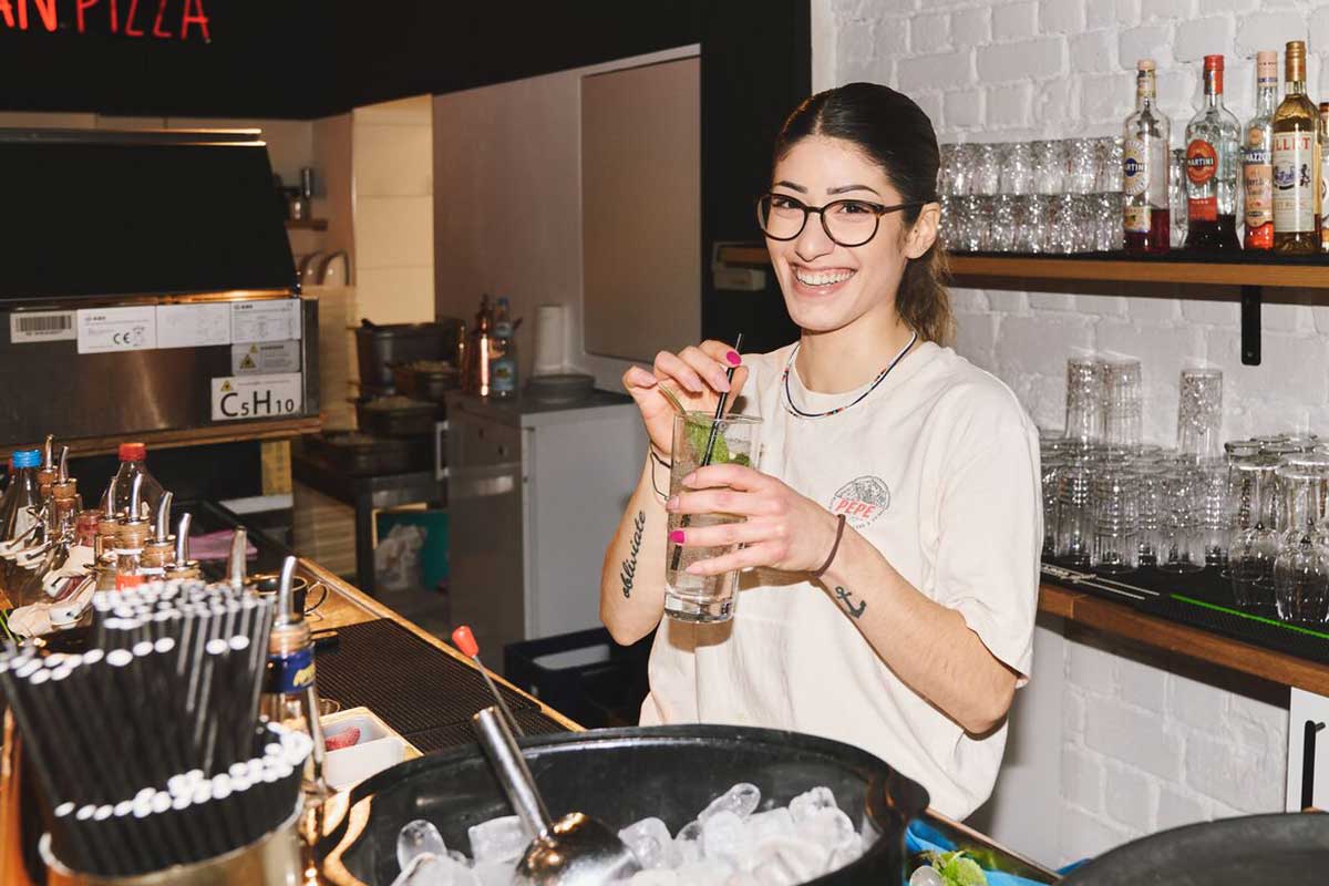 Frau mit braunen Haaren und Brille steht mit einem Drink in der Hand hinter einer Bar" title="Barkeeperin Pepe im Cosmo