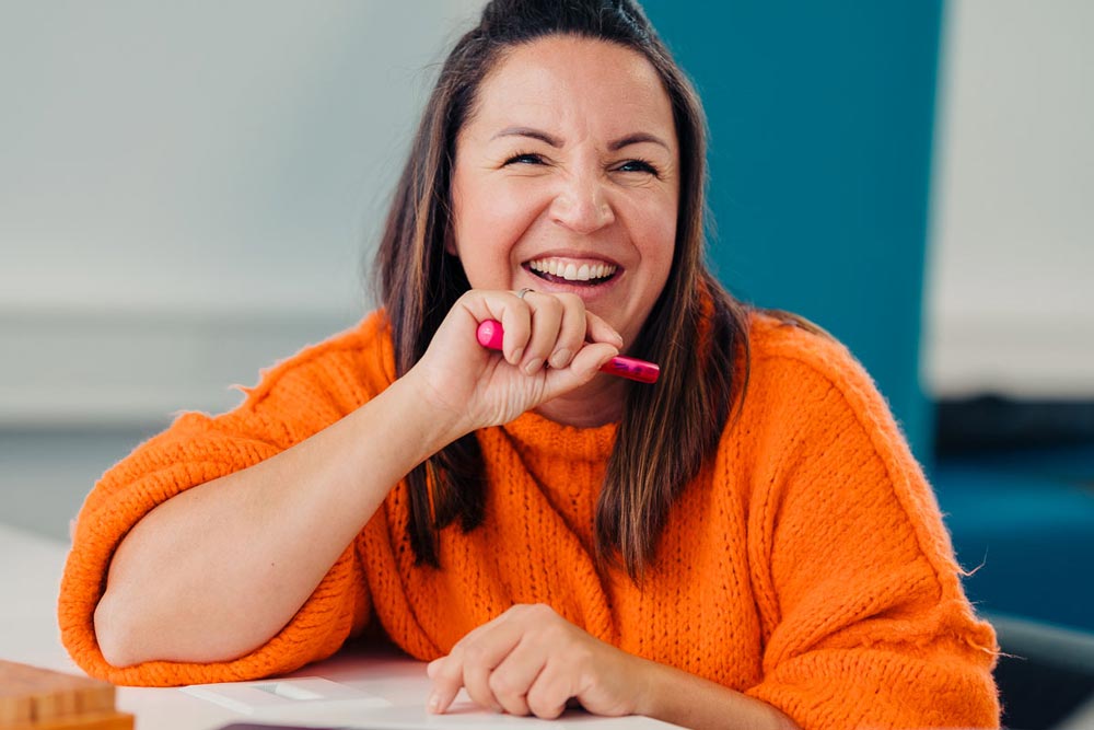 eine lachende Frau mit Stift in der Hand sitzt am Schreibtisch" title="Conny mit Stift am Schreibtisch