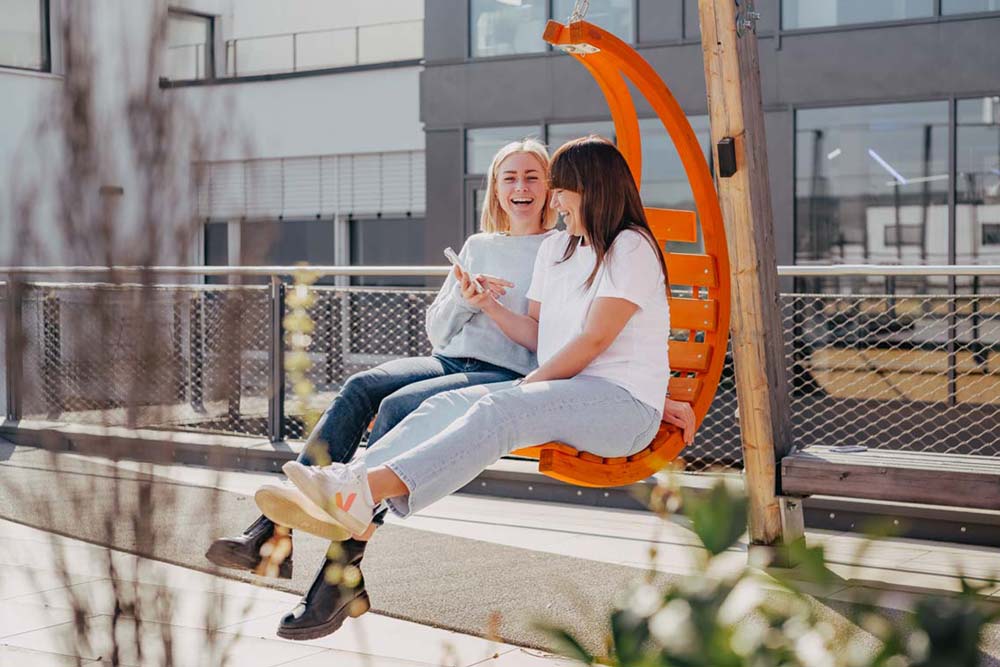 zwei Frauen sitzen lachend auf einem orangenen Hängestuhl auf einer Terrasse" title="Luise und Ilka im Hängestuhl