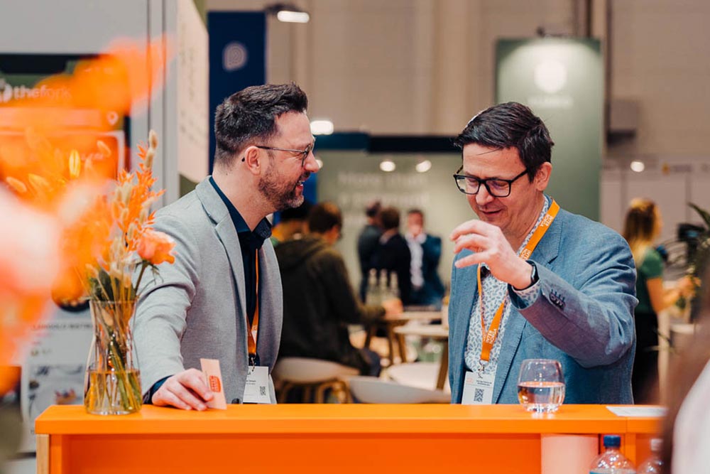 zwei Männer in Anzug stehen an einem Messestand und unterhalten sich fröhlich." title="Sebastian in einem Kundengespräch auf der Internorga