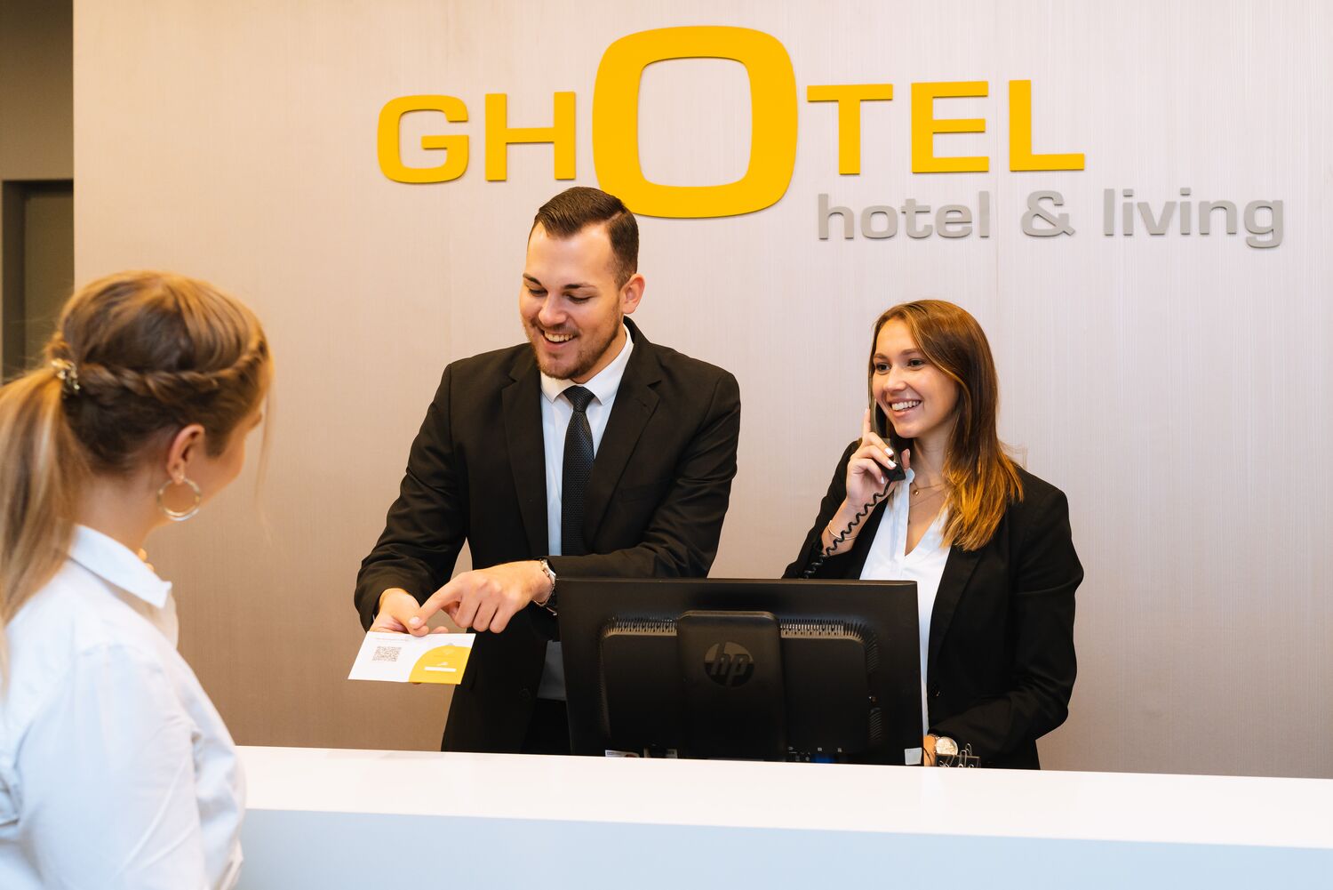 Mann steht neben Telefonistin an Rezeption und zeigt Frau etwas auf einer Zimmerkarte beim GHotel" title="branchen-hotellerie-header-ghotel-e2n
