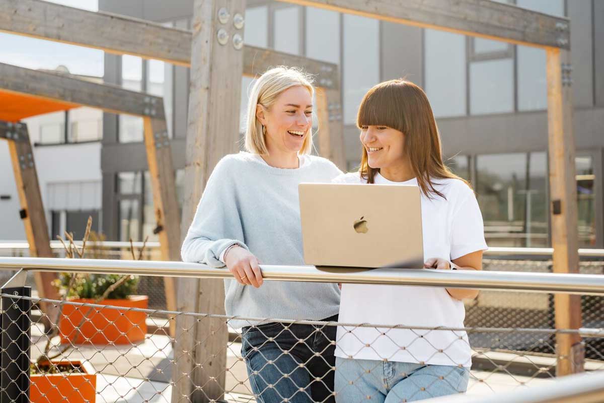 Zwei lachende Frauen stehen an einem Geländer mit einem Laptop in der Hand." title="Ilka und Luise beim Kundenservice