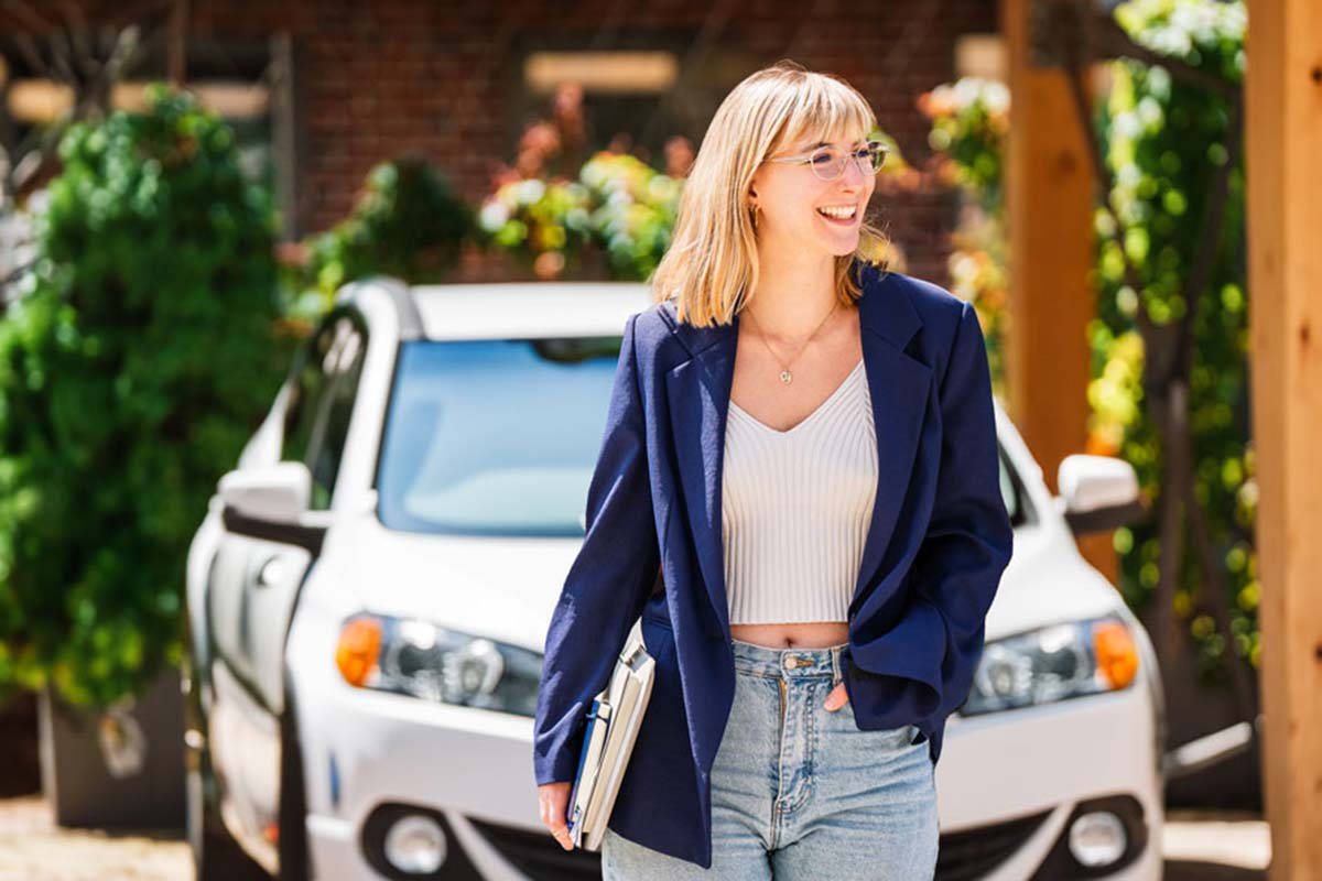 Eine lächelnde blonde Frau steht vor einem weißen Auto auf einem begrünten Parkdeck" title="Marie auf dem e2n Parkdeck