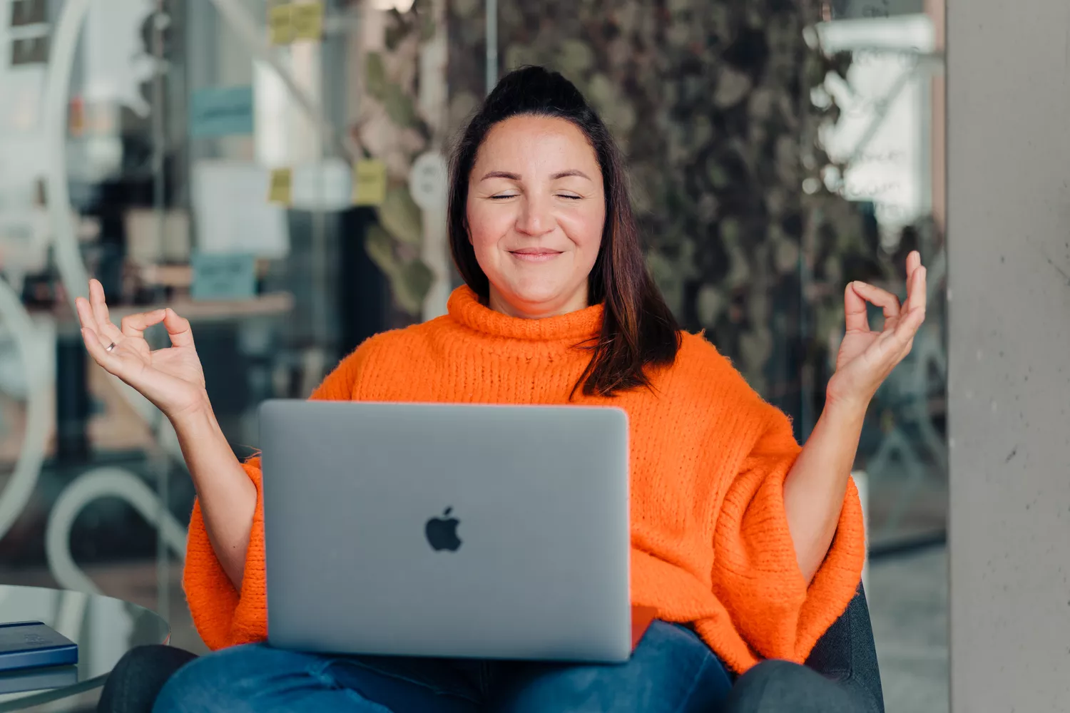 Frau in orangenem Pulli sitzt lächelnd mit geschlossenen Augen in Meditationshaltung auf einem Stuhl mit ihrem Laptop auf dem Schoß
