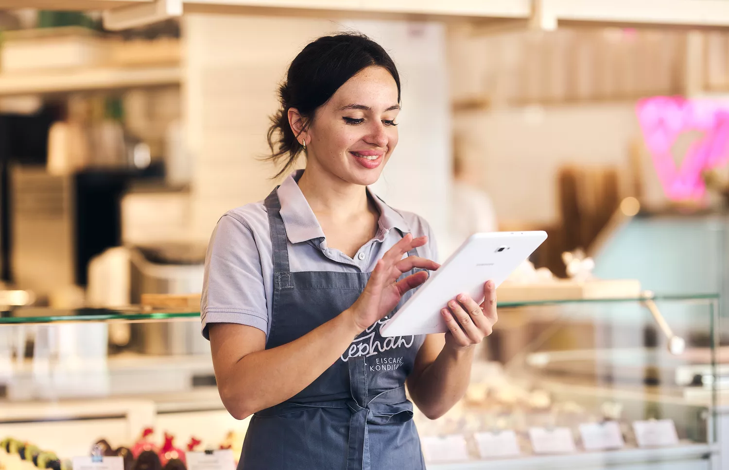 Brunette Kellnerin in Schürze schaut lachend in Cafe auf ihr Arbeitstablet und sticht sich zu ihrem Arbeitsbeginn ein" title="Sonntagszuschläge-e2n-header