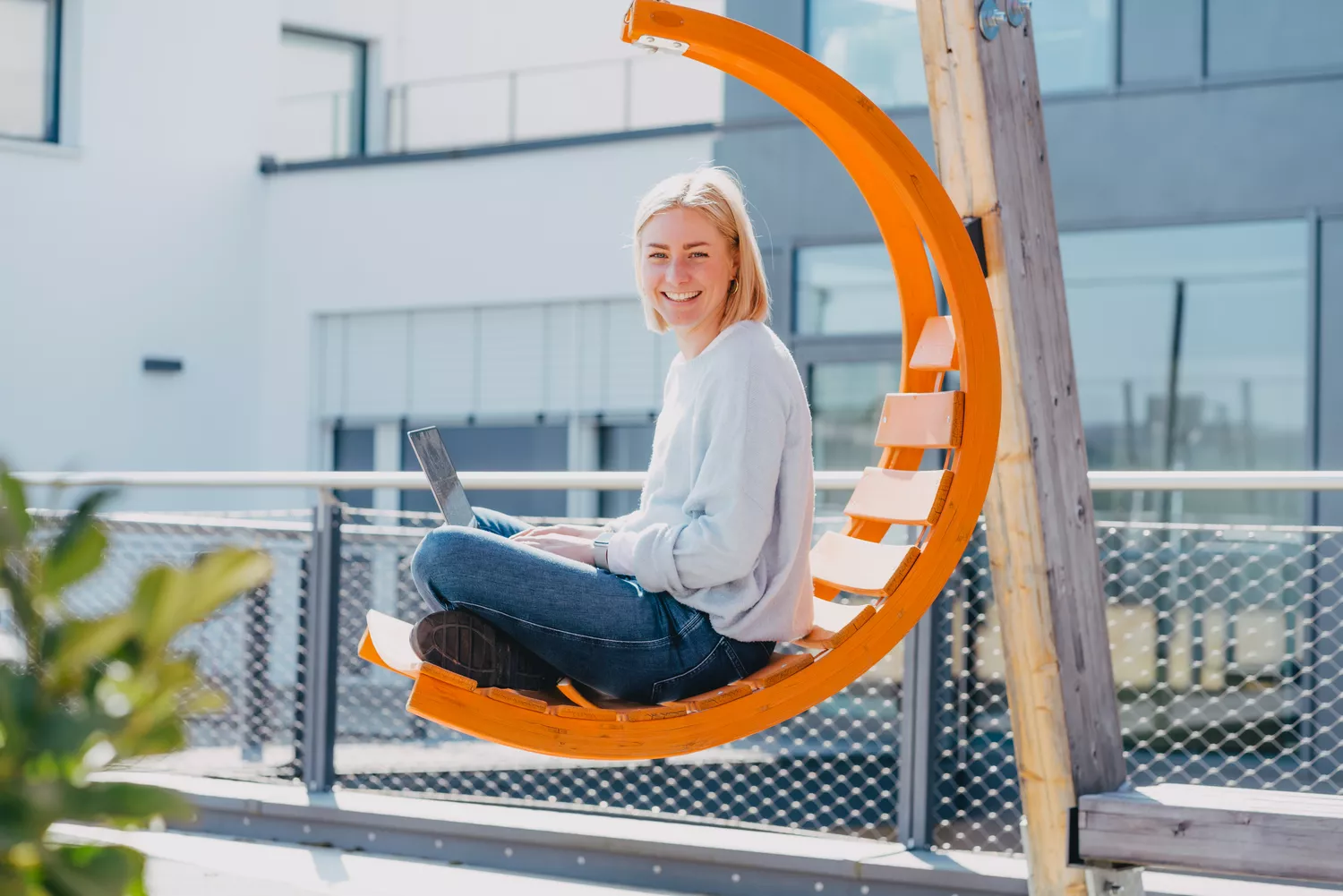 Blonde Frau in blauem Pulli sitzt draußen lachend auf orangenem Schaukelstuhl mit einem Laptop, der die e2n Software zeigt, auf dem Schoß" title="Teilzeit-e2n-zeiterfassung