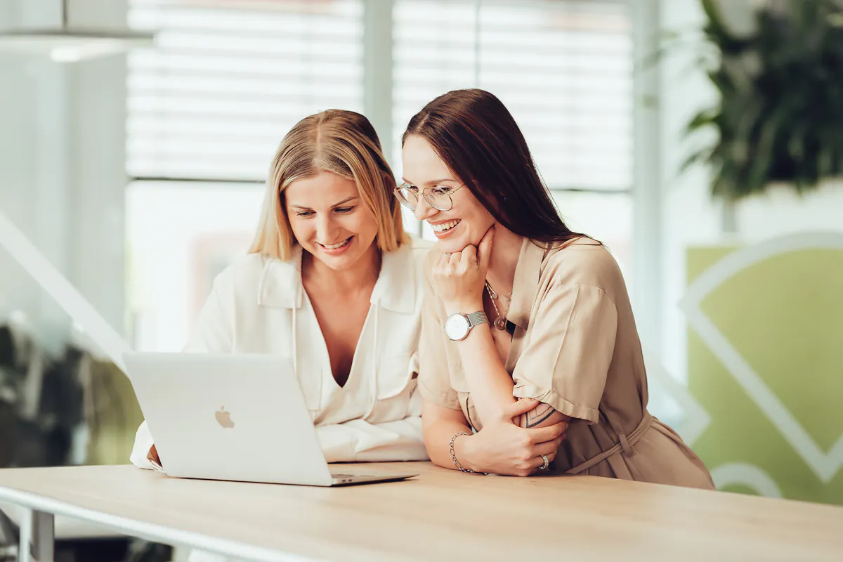 zwei junge Frauen lehnen auf einen Tisch und schauen lächelnd in ein Macbook" title="e2n-onboarding-pro-klein-ipad