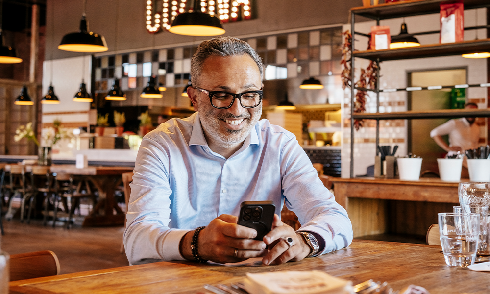 Nima Sabouni sitzt lächelnd mit dem Smartphone in der Hand" title="Blog-SuccessStory-LOsteria-Bild1