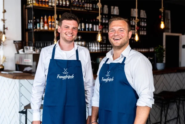 zwei Männer lächeln und tragen blaue Schürzen mit dem Logo von Fangfrisch drauf