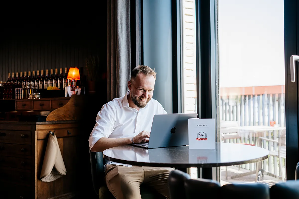 Ein Mann sitzt am Tisch in einem Restaurant und schaut lächelnd auf seinen Laptop